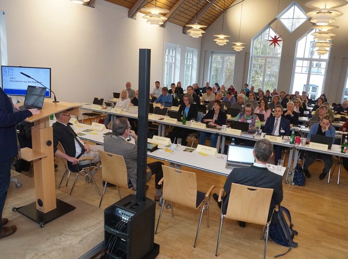 Eine Frau am rechten Rand des Bildes steht am Rednerpult und schaut in eine Gruppe an Menschen, die an Tischen sitzen.