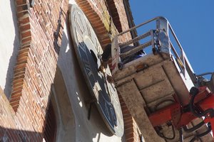 Eine Gondel eines Hubsteigers am Kirchturm. Ein Mann arbeitet am Ziffernblatt der Kirchturmuhr.