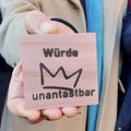 Eine Hand hält ein Holzstück mit der Aufschrift "Würde unantastbar" und einer Krone darauf