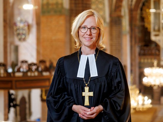 Bischöfin Nora Steen steht im Talar mit Bischofskreuz im Schleswiger Dom, im Hintergrund ist unscharf die Orgel